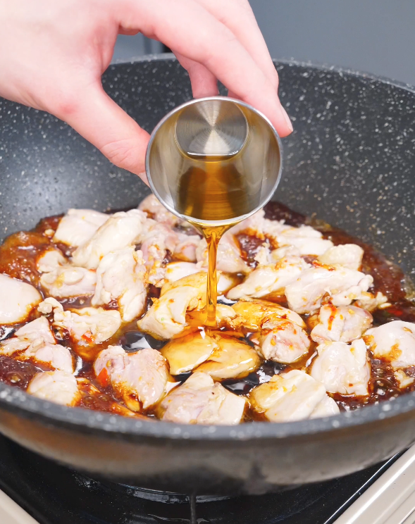 Sesame oil being poured into the pan with chicken and the soy-based sauce while making this Three Cup Chicken Recipe.