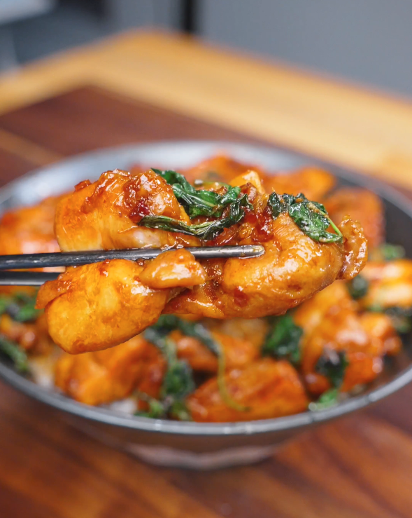 Chopsticks holding a piece of Three Cup Chicken coated in a glossy soy-based sauce with wilted Thai basil.
