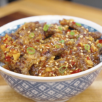 A bowl of Crispy Chili Beef Recipe served over rice and topped with sesame seeds, scallions, and a sticky sweet chili garlic sauce.