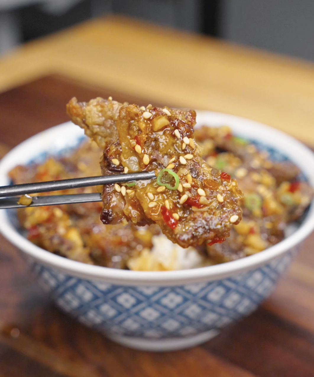 A piece of Crispy Chili Beef Recipe being held with chopsticks, showing the crispy coating covered in sweet chili garlic sauce and topped with sesame seeds and scallions.
