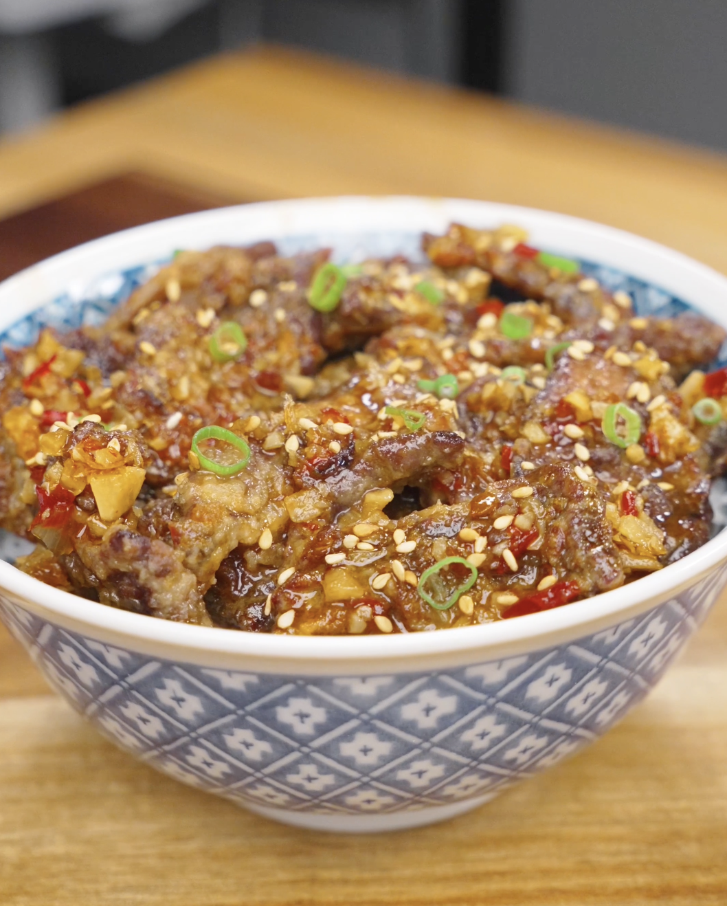 A bowl of Crispy Chili Beef Recipe served over rice and topped with sesame seeds, scallions, and a sticky sweet chili garlic sauce.