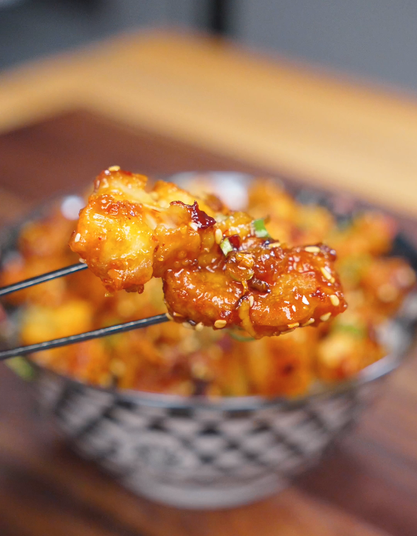 A close-up of a crispy piece of Crispy Chili Oil Chicken coated in a sticky chili oil sauce and held with chopsticks.