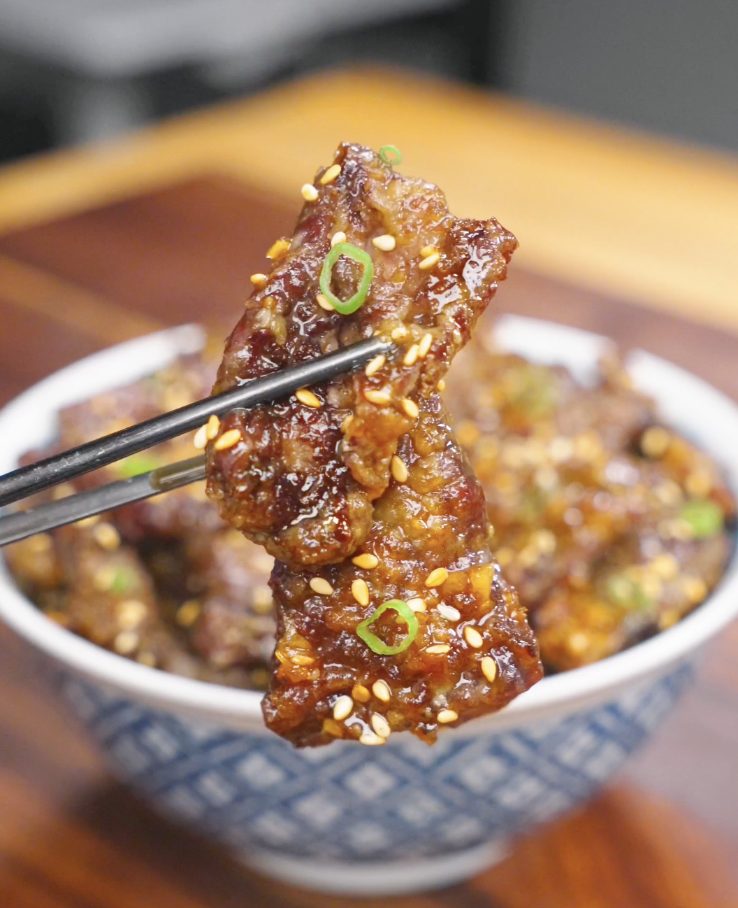 Piece of Crispy Honey Garlic Beef Recipe held with chopsticks above a bowl of crispy beef. The beef is coated in a sticky honey garlic glaze and finished with sesame seeds and fresh scallions.