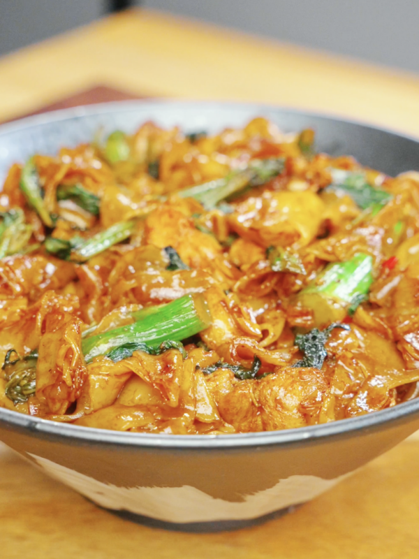 Bowl of Drunken Noodles Recipe made with wide rice noodles, chicken thighs, Chinese broccoli, and Thai basil. The noodles are coated in a rich sauce and garnished with fresh vegetables.