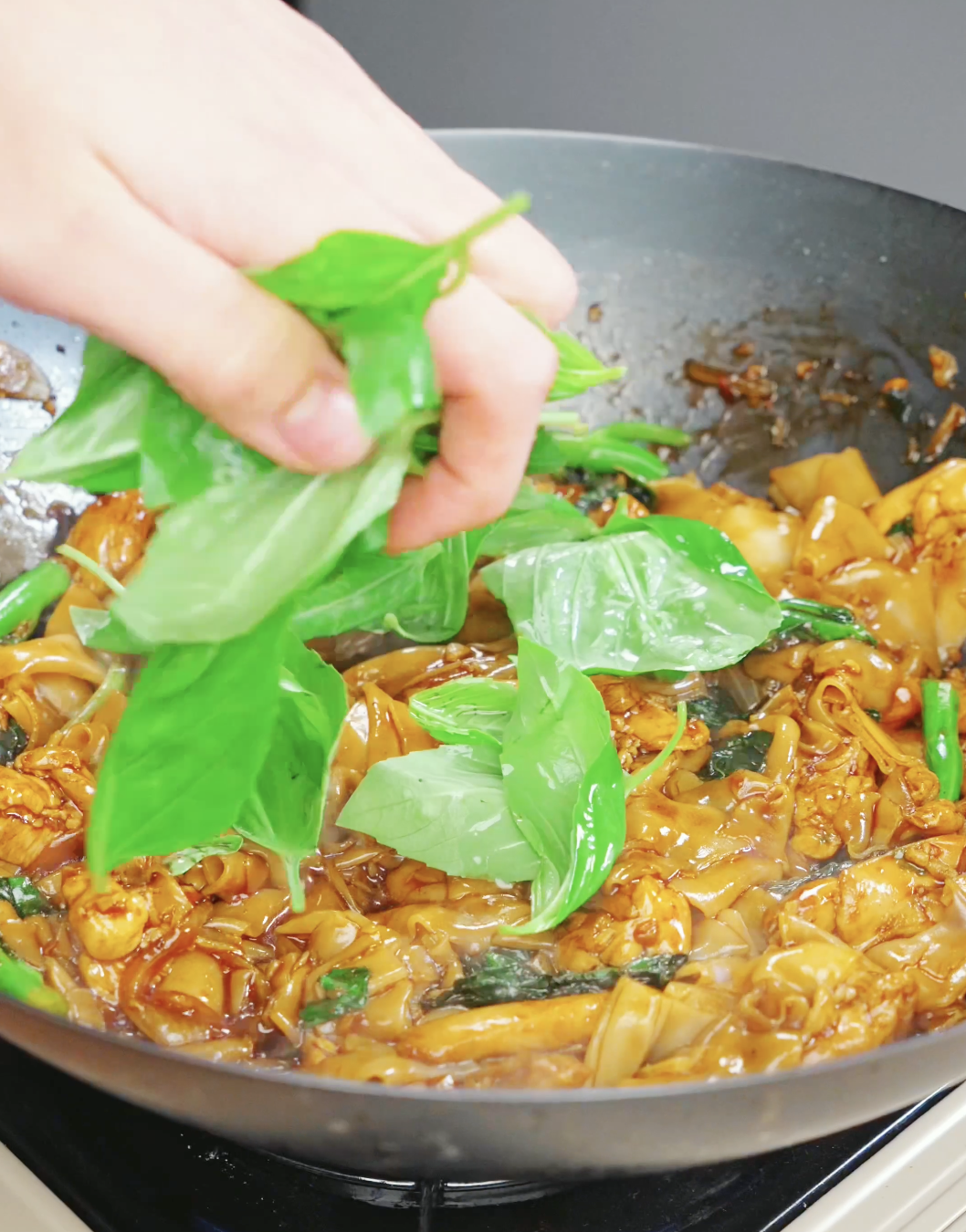 Fresh Thai basil leaves being added to a wok full of Drunken Noodles Recipe. The basil wilts into the noodles and adds the signature aroma that makes Pad Kee Mao so distinctive.