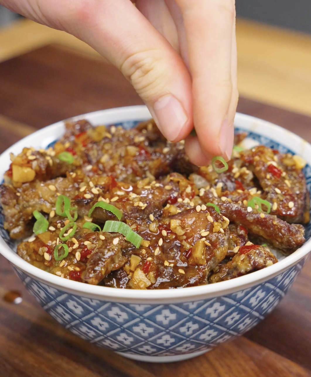 Fresh scallions being sprinkled over a bowl of Crispy Chili Beef Recipe finished with sesame seeds and sweet chili garlic sauce.