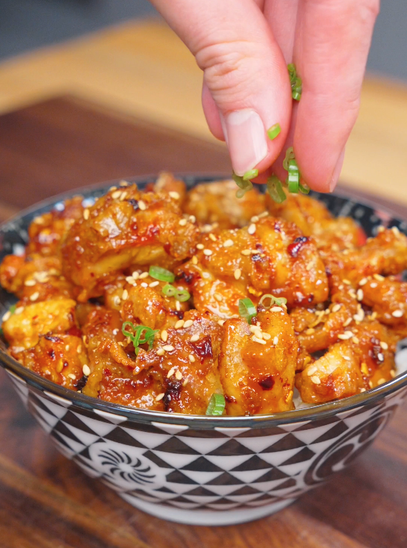 Fresh sliced scallions being sprinkled over a bowl of Crispy Chili Oil Chicken finished with sesame seeds.
