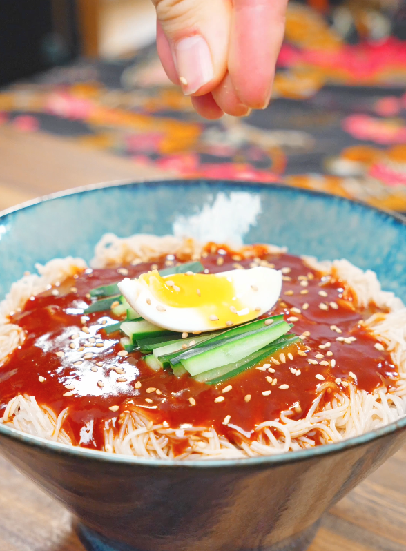 Sesame seeds being sprinkled over Spicy Cold Noodles Recipe topped with cucumber and a boiled egg.