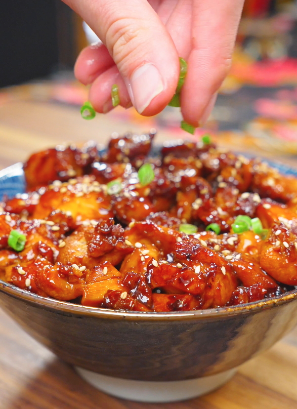 Fresh sliced scallions being sprinkled over a bowl of Sweet Honey Chicken Recipe finished with sesame seeds and served over steamed rice.