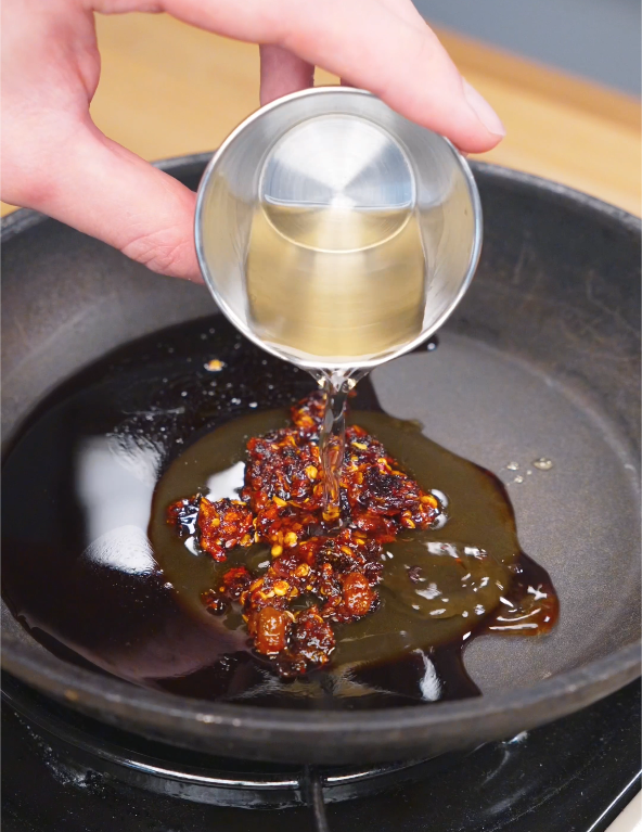 Rice vinegar being poured into a pan with soy sauce, honey, chili crisp, and garlic to make the sauce for Crispy Chili Oil Chicken.