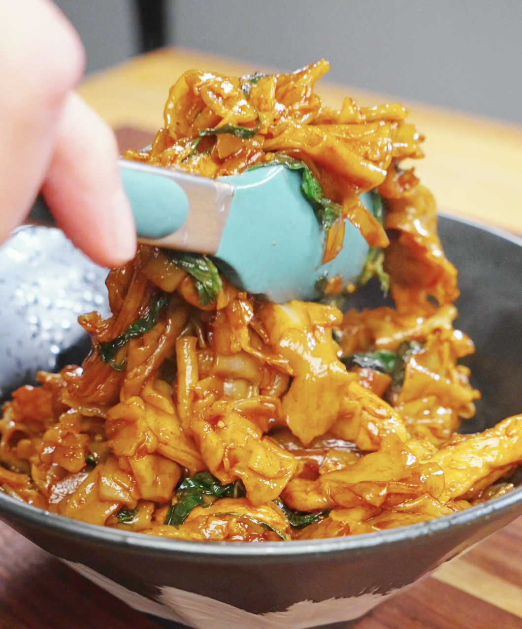 Freshly cooked Drunken Noodles Recipe being placed into a serving bowl. The wide rice noodles are coated in a savory sauce and tossed with chicken, Chinese broccoli, onions, chilies, and Thai basil.