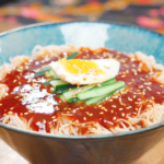 Spicy Cold Noodles Recipe served in a bowl with gochujang sauce, cucumber, boiled egg, and sesame seeds.