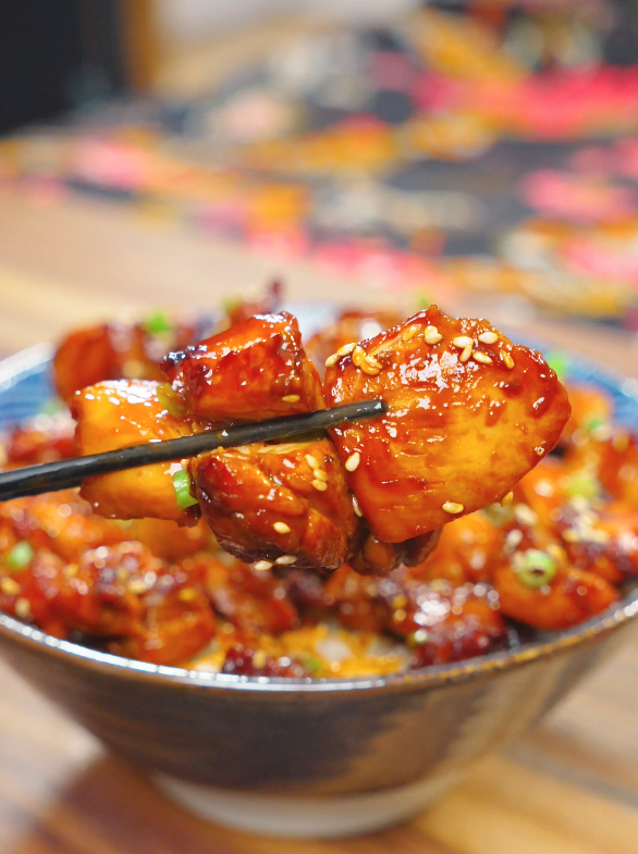 A close-up of a glazed piece from this Sweet Honey Chicken Recipe held with chopsticks, coated in a sticky honey glaze and sprinkled with sesame seeds.