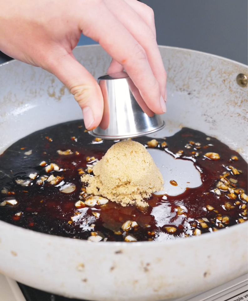 A scoop of brown sugar being added to a pan of garlicky soy sauce for Crispy Sticky Sweet Chicken Recipe.