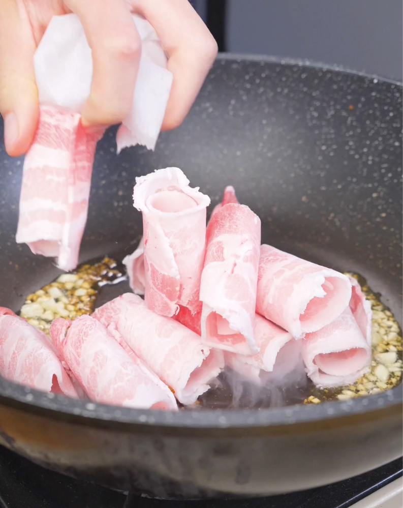 Rolled slices of pork belly being added to the garlic and oil in the pan for Sticky Sweet Pork Belly Recipe.