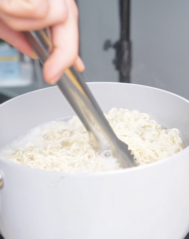 Instant ramen noodles being cooked and separated with tongs in boiling water for Hoisin Garlic Noodles Recipe.