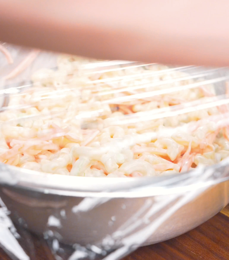 A bowl of dressed macaroni being covered with plastic wrap to chill for Hawaiian Mac Salad Recipe.
