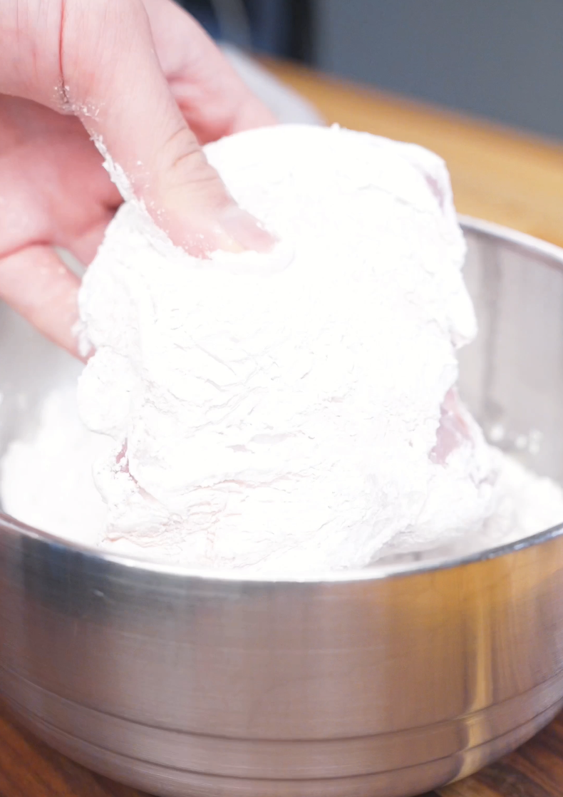 Chicken being tossed and coated in a bowl of starch while preparing Crispy Sticky Sweet Chicken Recipe.