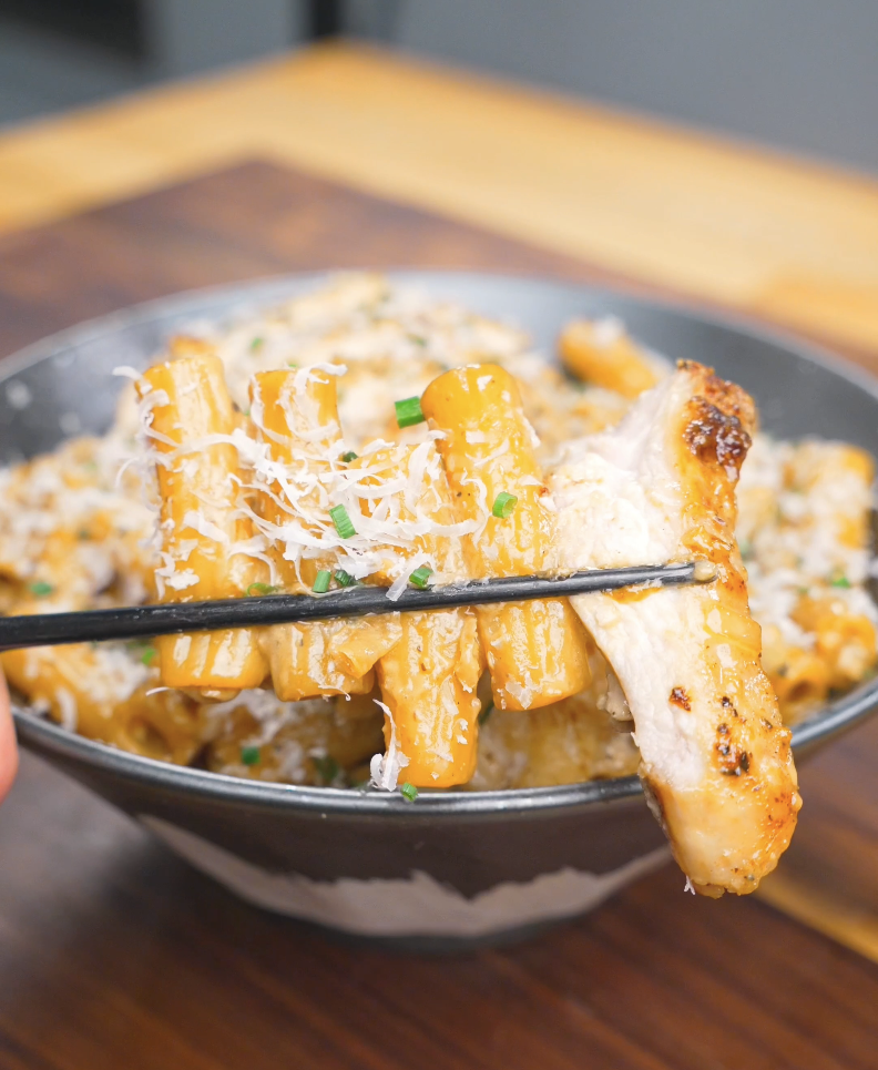 Chopsticks lifting a piece of chicken and saucy rigatoni above the bowl of Creamy Garlic Chicken Pasta Recipe.