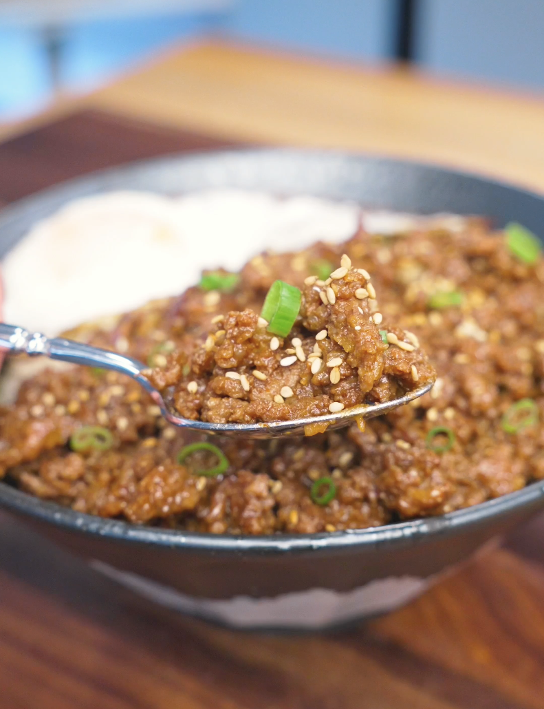 A spoonful of saucy, sesame-topped beef lifted above the bowl of Garlic Ground Beef Recipe.
