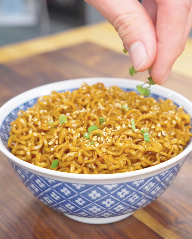 Sliced scallions being sprinkled over a sesame-topped bowl of Hoisin Garlic Noodles Recipe.