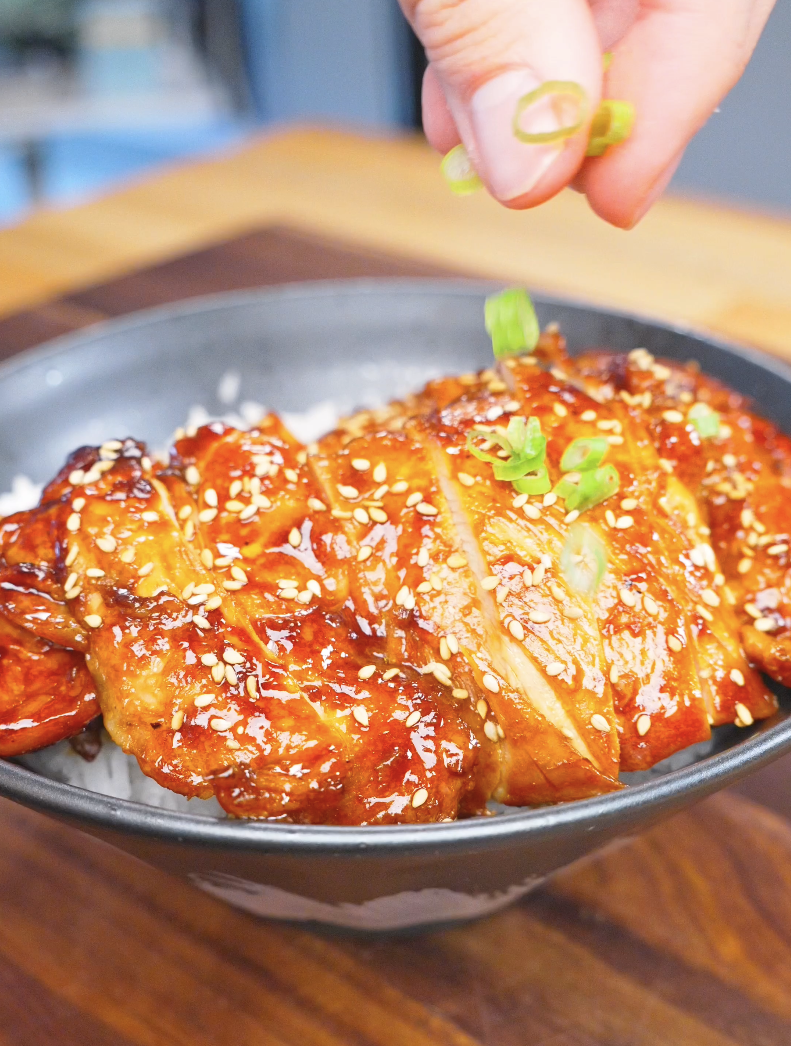Sliced green onions being sprinkled over a sesame-topped rice bowl of Huli Huli Chicken Recipe.