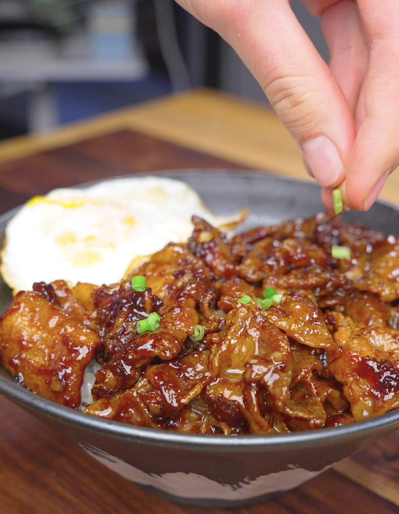 Sliced green onions being sprinkled by hand over a rice bowl of Sticky Sweet Pork Belly Recipe.