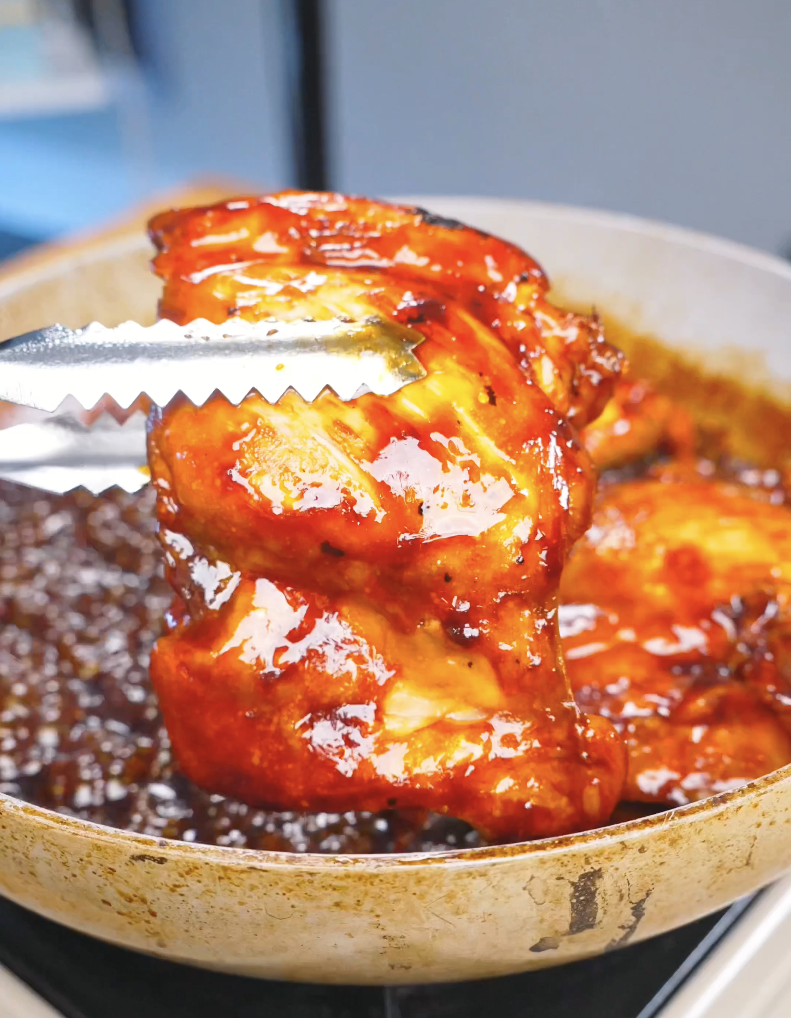 Tongs lifting chicken coated in a thick, glossy glaze from the pan while making Huli Huli Chicken Recipe.