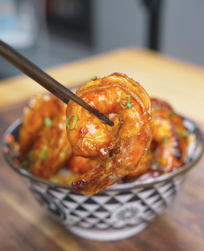 Chopsticks lifting a glossy glazed shrimp above the bowl of Honey Garlic Shrimp Recipe.