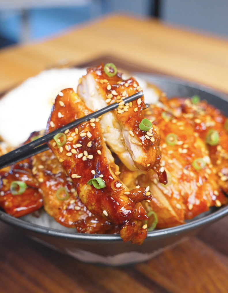 Chopsticks lifting a glazed, sesame-coated slice above the bowl of Huli Huli Chicken Recipe.