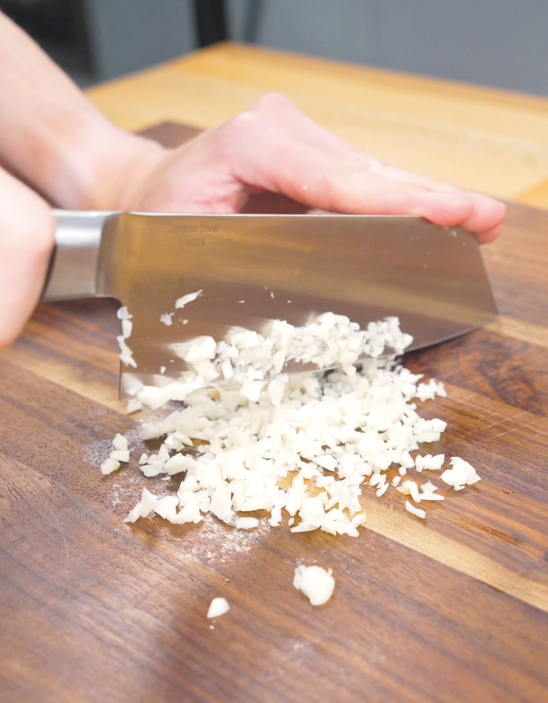 Fresh garlic being finely minced with a cleaver on a cutting board for Creamy Garlic Chicken Pasta Recipe.