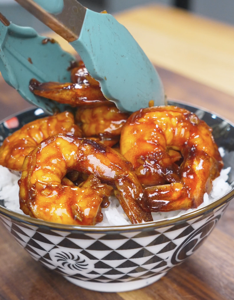 Glazed shrimp being placed over steamed rice with tongs for Honey Garlic Shrimp Recipe.