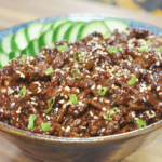 A bowl of Sticky Sweet Ground Beef Recipe served over rice with sliced cucumbers, sesame seeds, and scallions.