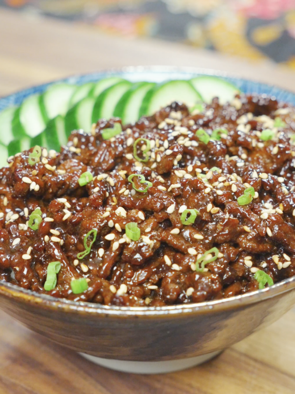 A bowl of Sticky Sweet Ground Beef Recipe served over rice with sliced cucumbers, sesame seeds, and scallions.