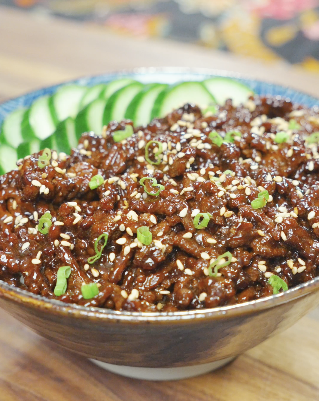 A bowl of Sticky Sweet Ground Beef Recipe served over rice with sliced cucumbers, sesame seeds, and scallions.