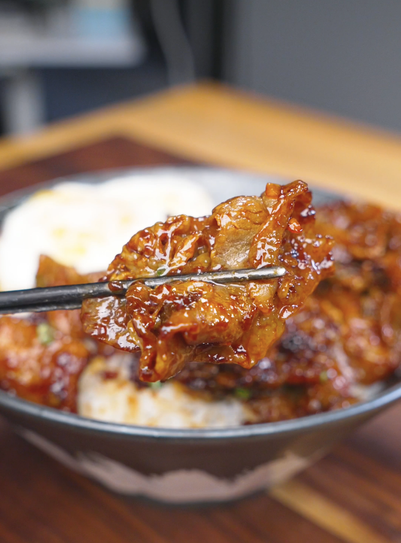 Chopsticks lifting a glossy, caramelized piece above the bowl of Sticky Sweet Pork Belly Recipe.
