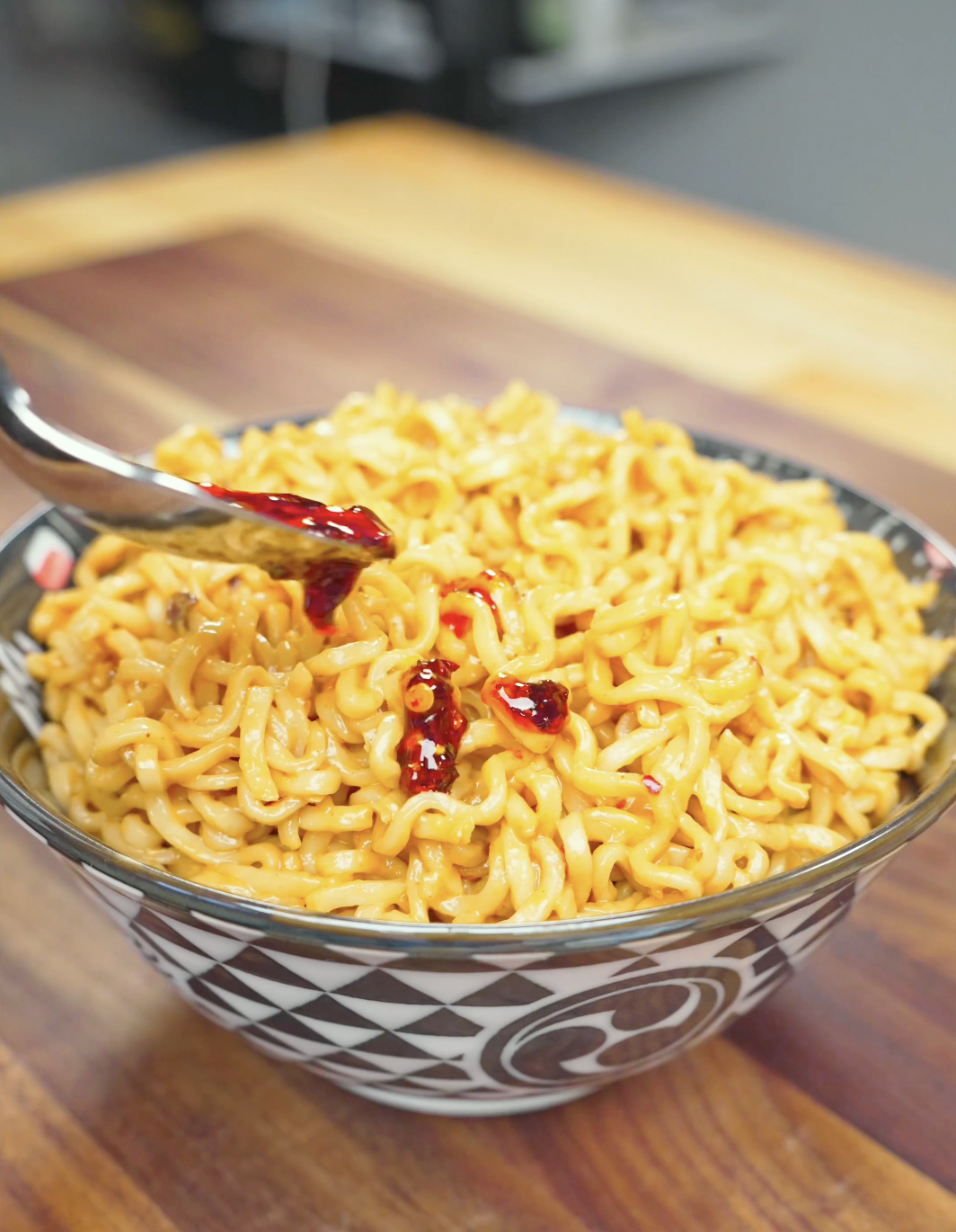 A spoonful of red chili oil being drizzled over a finished bowl of Spicy Peanut Noodles Recipe.