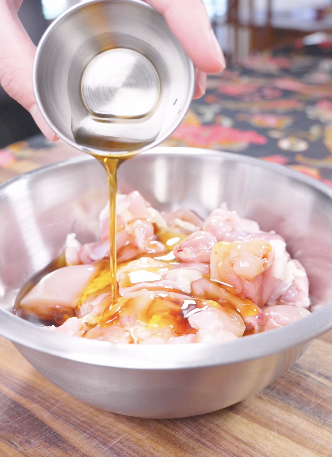 Golden sesame oil being poured over raw chicken pieces in a metal bowl for Honey Sesame Chicken Recipe.