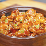 Finished Sticky Orange Chicken Recipe in a clay pot topped with sesame seeds and sliced scallions.