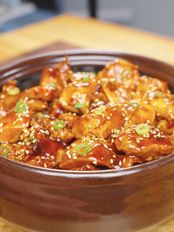 Finished Sticky Orange Chicken Recipe in a clay pot topped with sesame seeds and sliced scallions.