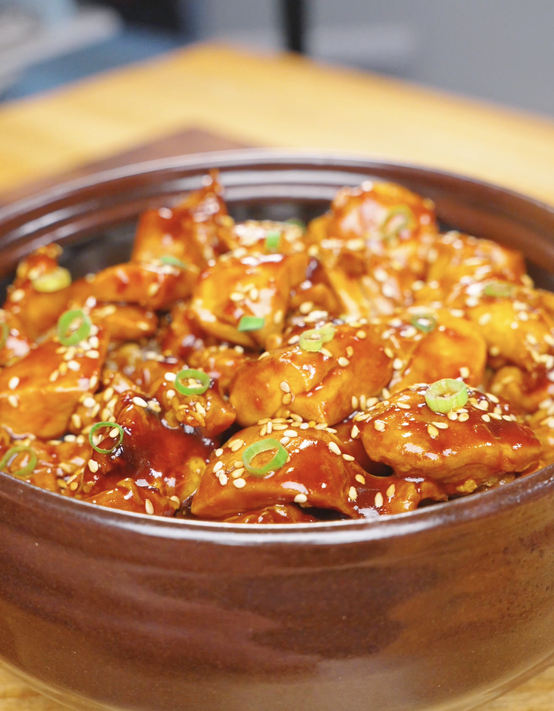 Finished Sticky Orange Chicken Recipe in a clay pot topped with sesame seeds and sliced scallions.