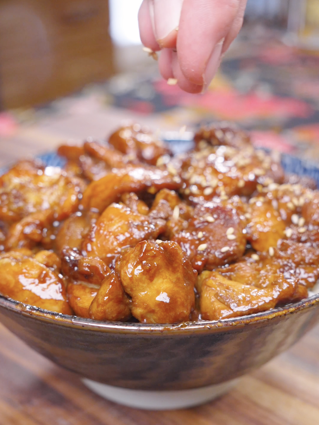 Sesame seeds being sprinkled by hand over a bowl of glazed Honey Sesame Chicken Recipe.