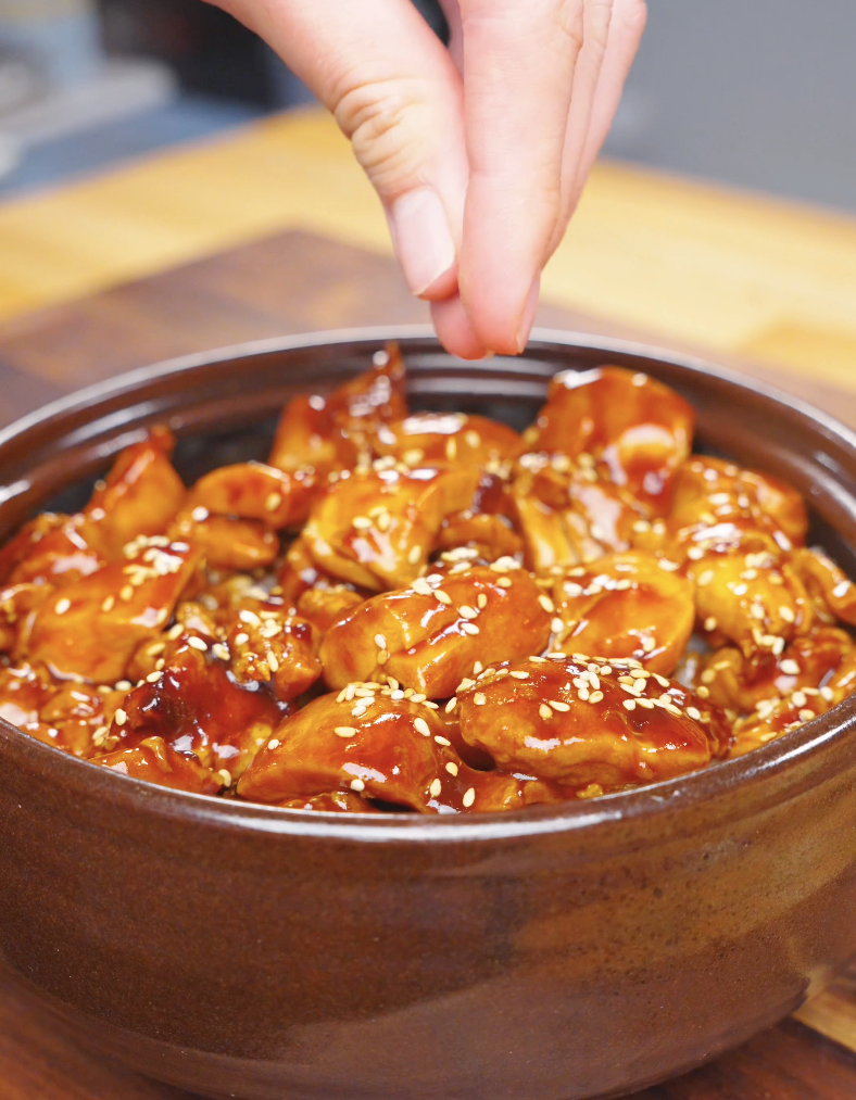 Sesame seeds being sprinkled over a clay pot of Sticky Orange Chicken Recipe.