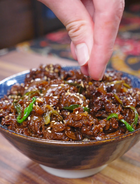 Sesame seeds being sprinkled over a ceramic bowl of Teriyaki Beef Recipe.