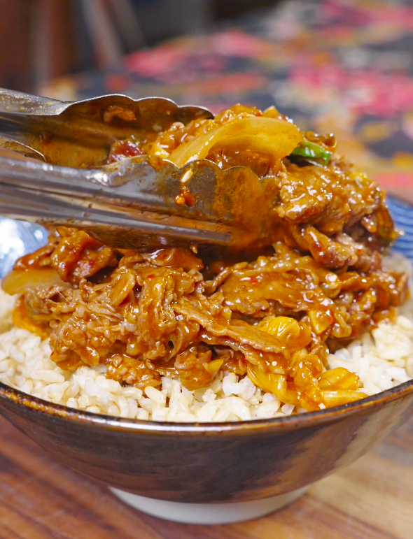 Saucy beef and kimchi being placed over steamed rice with tongs for Kimchi Beef Stir Fry Recipe.