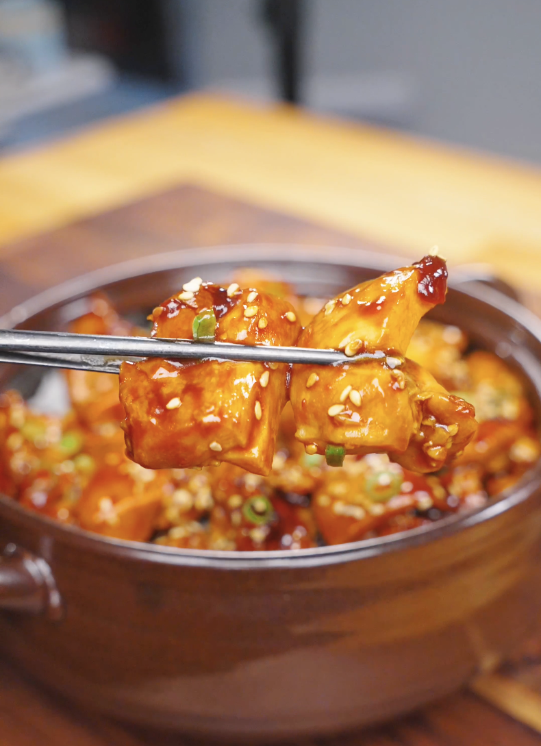 Chopsticks lifting glossy glazed chicken chunks above the bowl of Sticky Orange Chicken Recipe.