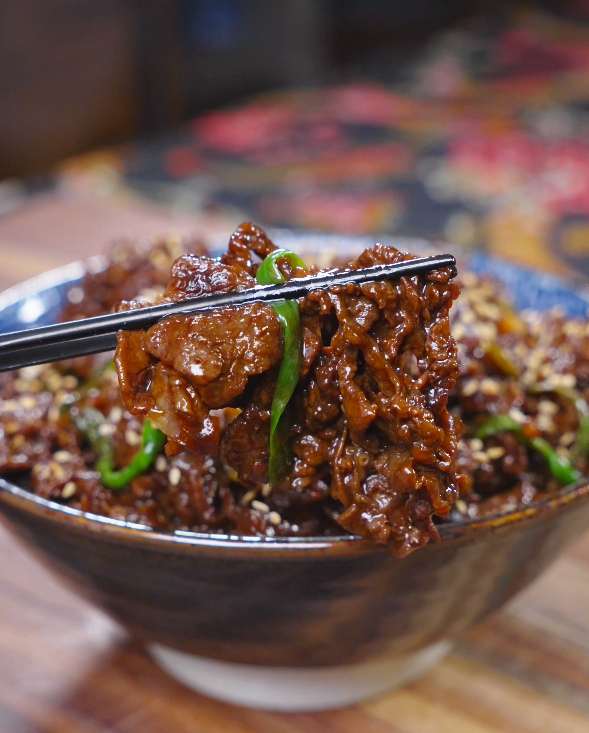 Chopsticks lifting a glossy, sesame-topped slice of beef above the bowl of Teriyaki Beef Recipe.