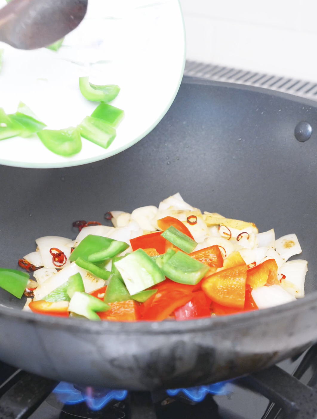 Green and red bell pepper chunks being tipped into the wok with the onions for Kung Pao Chicken Noodles Recipe.