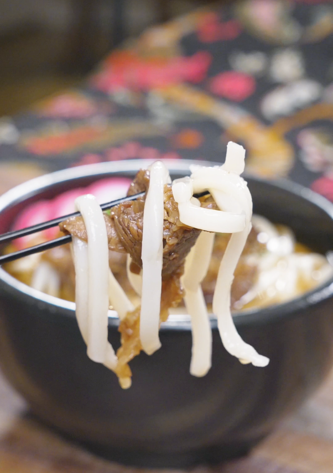 Chopsticks lifting chewy noodles and a piece of glazed beef above the bowl of Beef Udon Recipe.