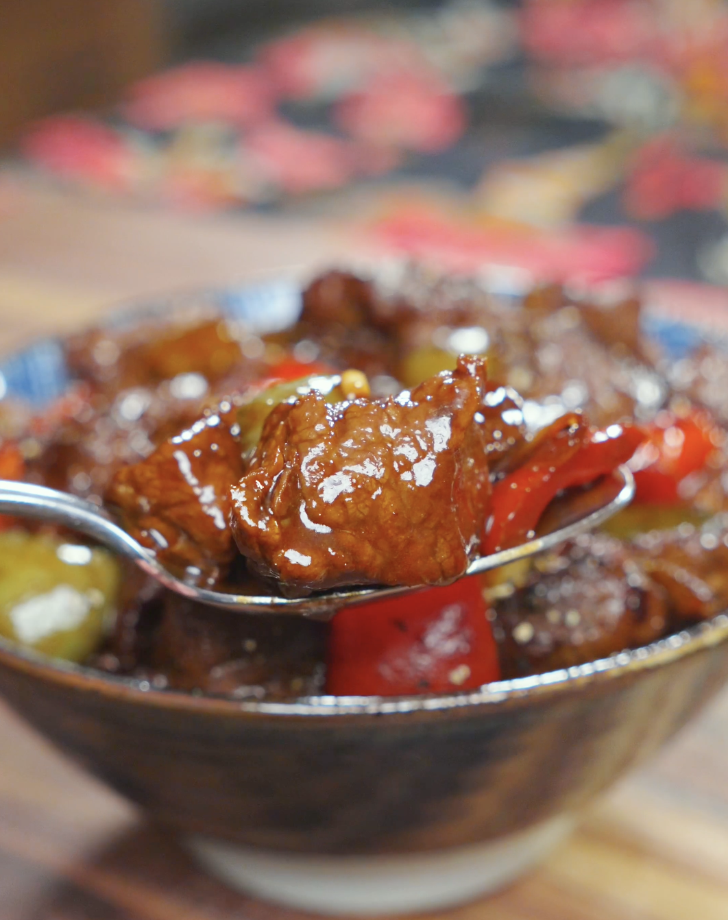 A spoon lifting a glossy steak cube and red pepper above the bowl of Black Pepper Steak Bites Recipe.